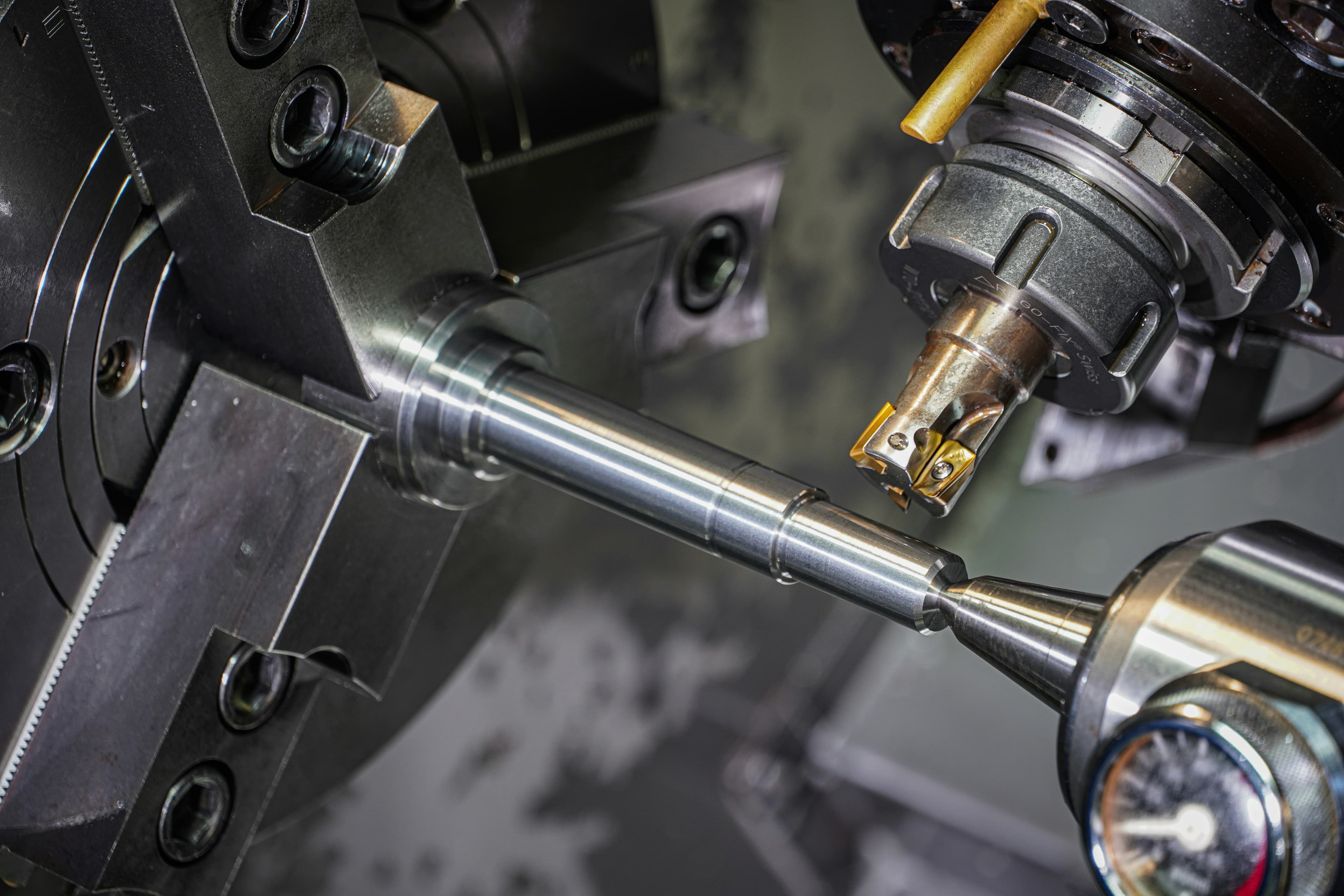 A precision lathe turning a stainless shaft, viewed close-up between the chuck and the live tool.