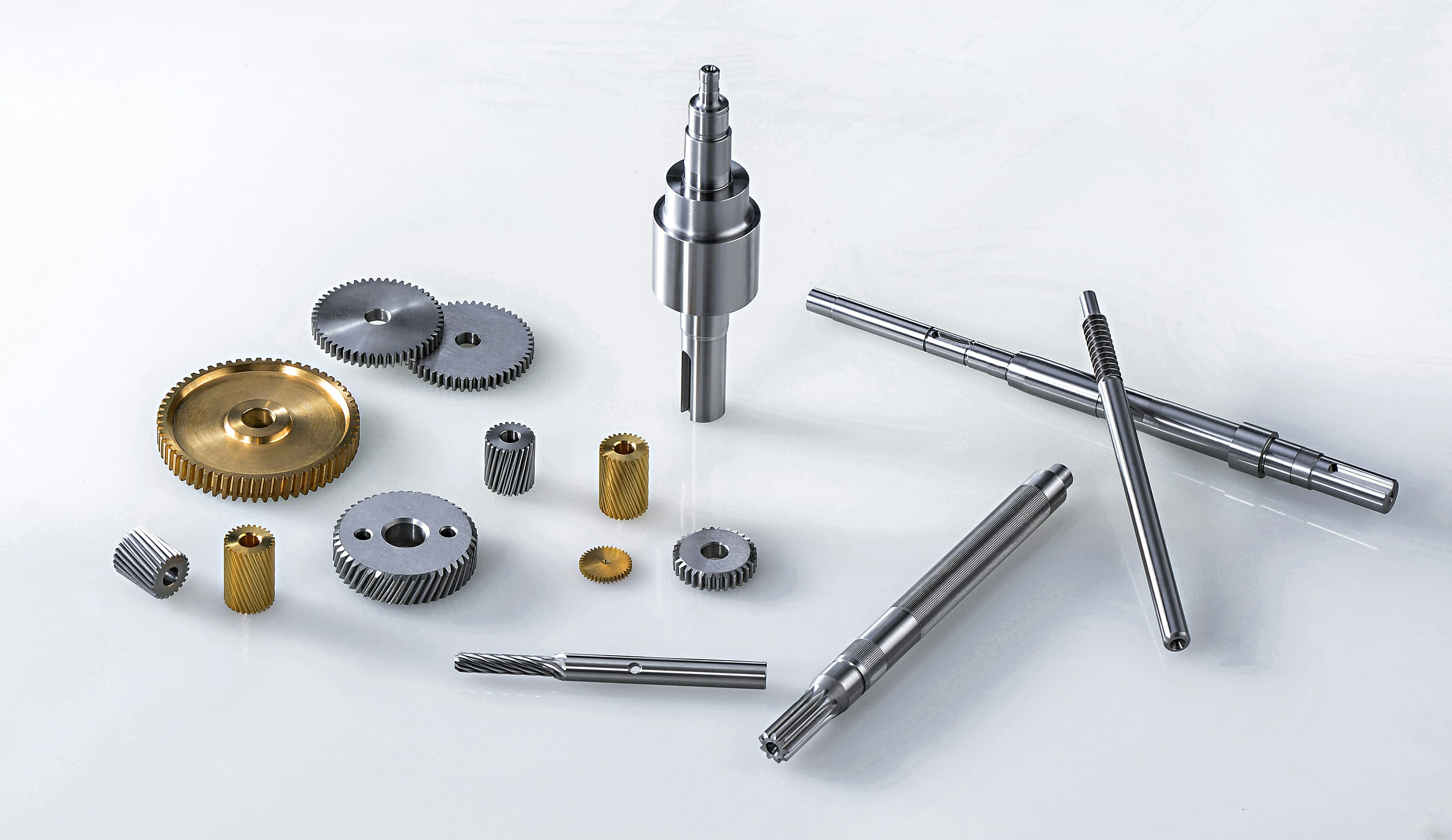 Flatlay of precision-machined parts — gears, shafts, bushings and a knurled tool — arranged on a soft white surface.